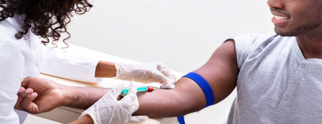 Young dark skin tone man in grey shirt having blood drawn by healthcare worker in medical gloves.