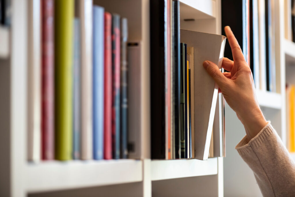 White bookshelf with colorful books, light skin tone hand pulling off-white book from shelf.